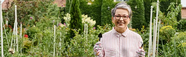 Jolly mature woman with glasses holding gardening tools in hands and smiling at camera, banner — Stock Photo