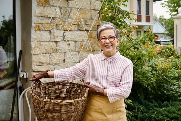 Cheerful mature woman holding big straw basket and smiling at camera near her house in England — Stock Photo