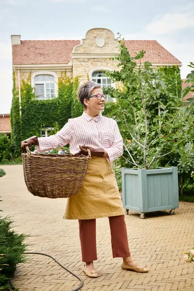 Веселая зрелая женщина в очках держит большую соломенную корзину и позирует возле своего дома в Англии — стоковое фото