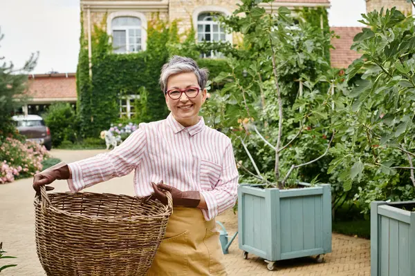 Весела зріла жінка тримає великий солом'яний кошик і посміхається на камеру біля свого будинку в Англії — стокове фото