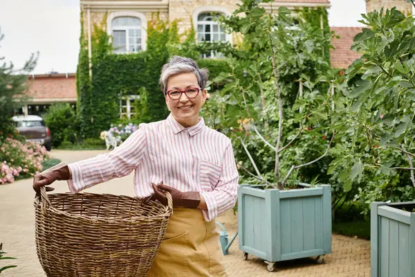 Mulher madura alegre segurando grande cesta de palha e sorrindo para a câmera perto de sua casa na Inglaterra — Fotografia de Stock