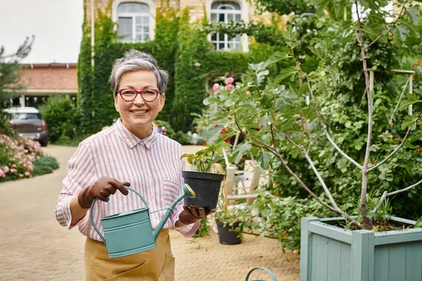 Appealing joyful mature woman holding watering can and pot with plant and smiling at camera, England — Stock Photo