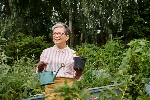 Beautiful cheerful mature woman holding watering can and pot with plant in her garden in England — Stock Photo