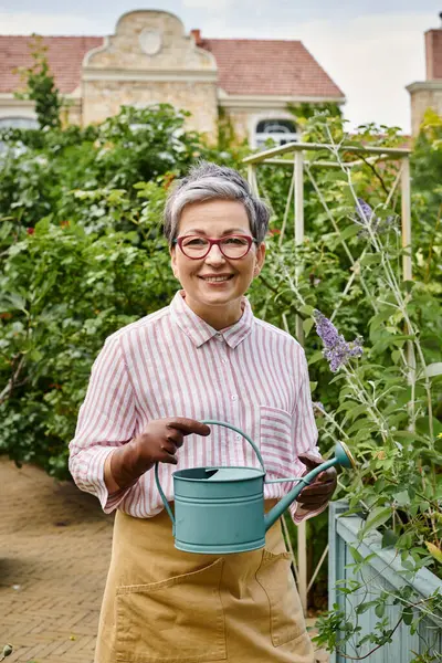 Alegre mujer madura con vasos sosteniendo regadera y sonriendo a la cámara cerca de casa en Inglaterra - foto de stock