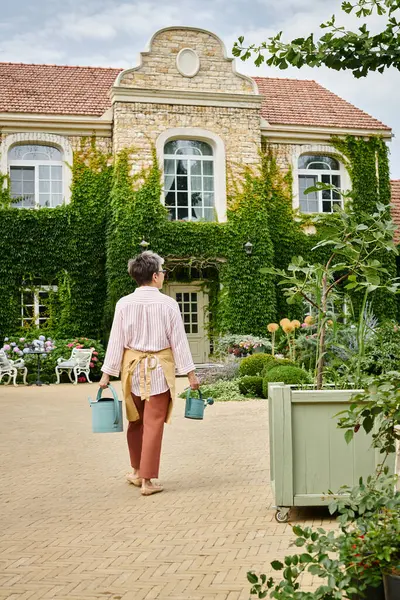 Back view of mature woman in cozy attire holding two watering cans in front of her house in England — Stock Photo