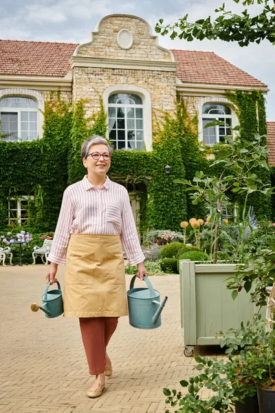 Attractive cheerful mature woman in casual attire holding watering cans in front of house in England — Stock Photo