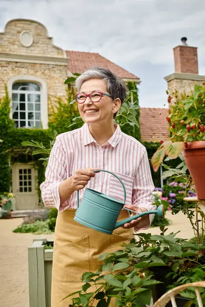 Добре виглядає радісна зріла жінка поливає свої живі яскраві квіти в її саду в Англії — стокове фото
