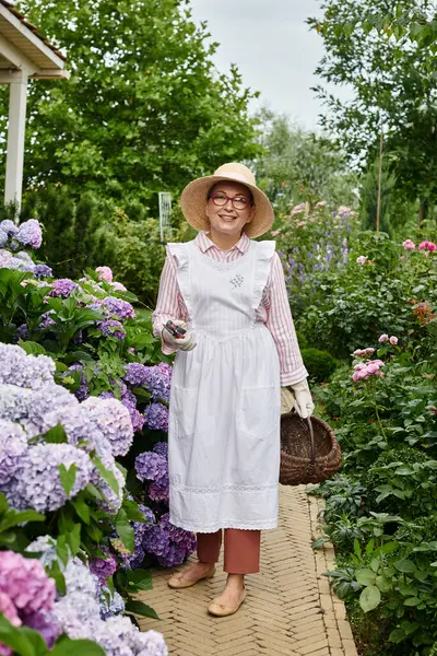 Schöne reife fröhliche Frau mit Hut und Brille, die neben Hortensien posiert und in die Kamera schaut — Stockfoto