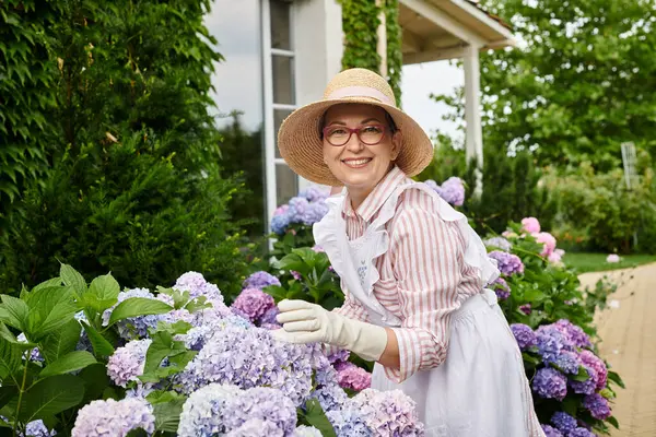 Attraktive, fröhliche reife Frau mit Hut, die sich um ihre Hortensie kümmert und in die Kamera lächelt — Stockfoto