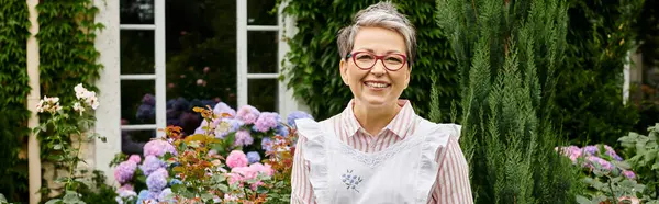 Mature cheerful woman with glasses posing next to flowers and smiling happily at camera, banner — Stock Photo