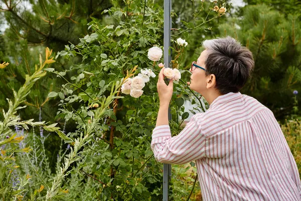 Atraente mulher alegre madura com óculos em trajes casuais cheirando rosas enquanto em seu jardim — Fotografia de Stock