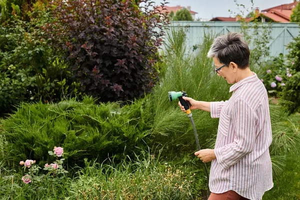 Belle femme mature gaie avec des lunettes en utilisant un tuyau pour arroser ses plantes vivantes dans son jardin — Photo de stock
