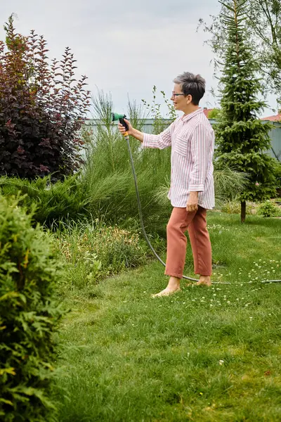 Bella cercando allegra donna matura con gli occhiali utilizzando tubo per innaffiare le sue piante vivaci nel suo giardino — Foto stock