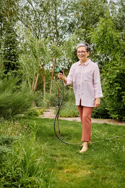 Boa aparência mulher madura alegre com óculos usando mangueira para regar suas plantas vivas em seu jardim — Fotografia de Stock