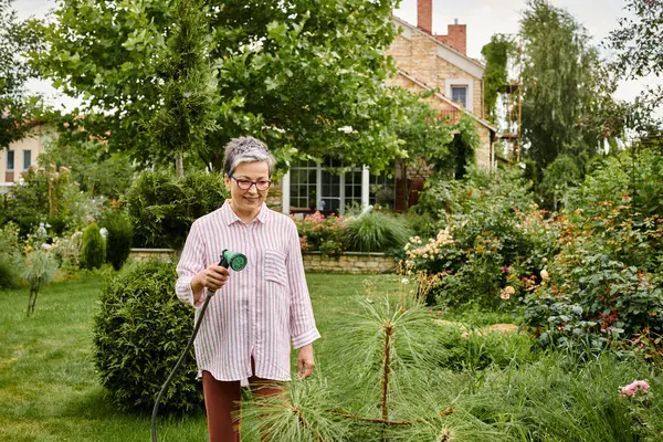 Bella cercando jolly donna matura con gli occhiali utilizzando tubo per innaffiare le sue piante vivaci nel suo giardino — Foto stock