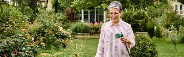 Hermosa mujer madura alegre con gafas usando manguera para regar plantas vivas en su jardín, pancarta - foto de stock