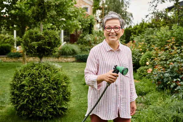 Atractiva mujer madura alegre en traje casual regando sus plantas vibrantes y sonriendo a la cámara - foto de stock
