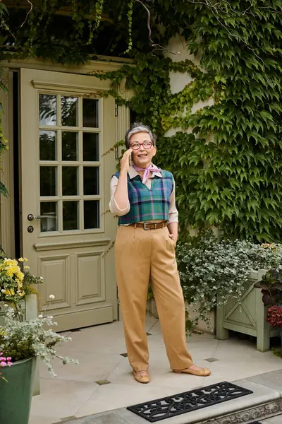Зрелая веселая элегантная женщина в очках и коротких волосах разговаривает по телефону рядом с домом в Англии — стоковое фото