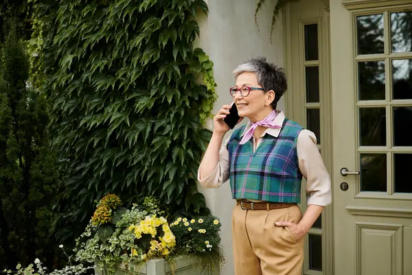 Mature cheerful elegant woman with glasses and short hair talking by her phone near house in England — Stock Photo