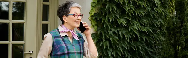 Зрелая радостная элегантная женщина в очках разговаривает по телефону рядом с домом в Англии, баннер — стоковое фото