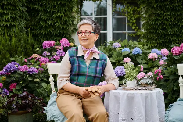 Hermosa mujer madura alegre sentado en el jardín a la hora del té cerca de la casa en Inglaterra y mirando hacia otro lado - foto de stock