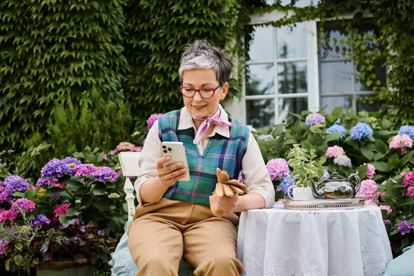 Reife, fröhliche Frau, die zur Teezeit im Garten sitzt und in der Nähe ihres Hauses in England auf das Telefon schaut — Stockfoto