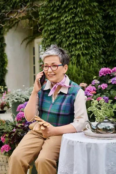 Mature cheerful woman sitting in garden at tea time and talking by phone near house in England — Stock Photo