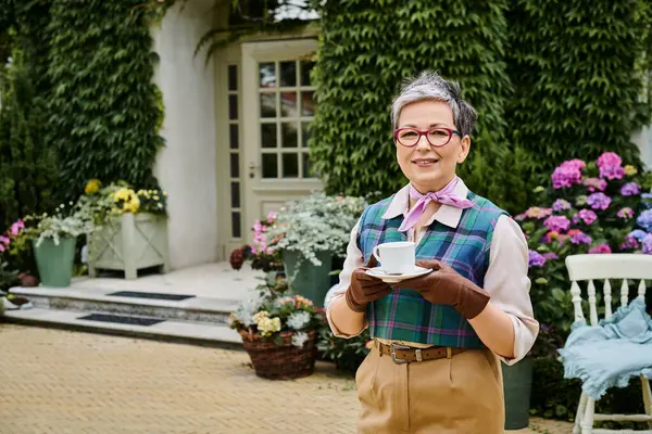 Elegant jolly mature woman in chic attire drinking tea near house in England and smiling at camera — Stock Photo
