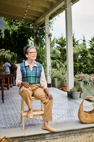 Модная веселая зрелая женщина сидит на стуле на крыльце своего дома в Англии и смотрит в сторону — стоковое фото
