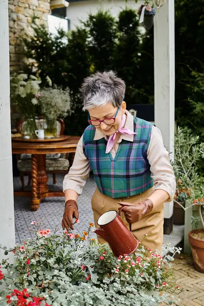 Attractive jolly mature woman in chic attire watering her flowers near her house in England — Stock Photo