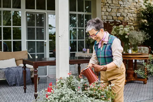 Attractive jolly mature woman in chic attire watering her flowers near her house in England — Stock Photo