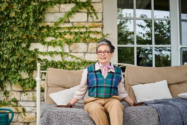Attractive joyous mature woman sitting on sofa near her house in England and looking at camera — Stock Photo