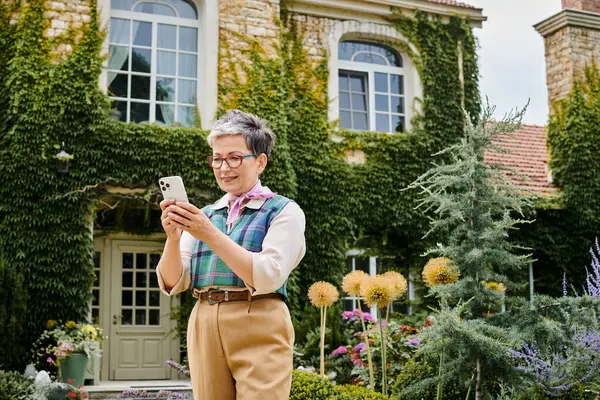 Mujer madura alegre sofisticada en traje elegante mirando su teléfono inteligente cerca de su casa en Inglaterra - foto de stock