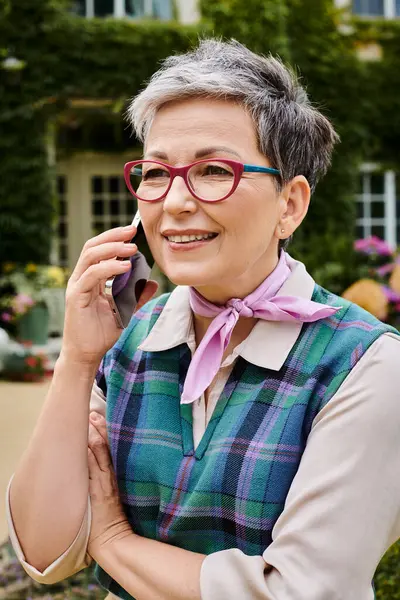 Элегантная веселая, зрелая женщина в стильных очках разговаривает по телефону по сети со своим домом в Англии — стоковое фото