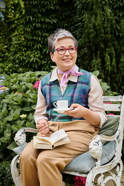 Mujer alegre madura refinada con vasos disfrutando de té caliente y libro cerca de su casa en Inglaterra - foto de stock