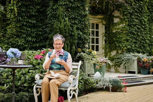 Изысканная зрелая веселая женщина с очками, наслаждающаяся горячим чаем и книгой возле своего дома в Англии — стоковое фото