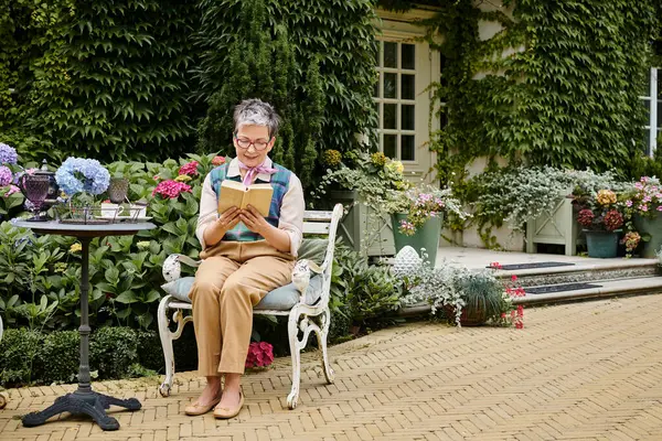 Elegant mature cheerful woman with glasses reading book at tea time near her house in rural England — Stock Photo