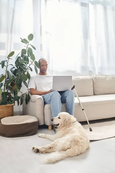 Un uomo afroamericano disabile con miastenia gravis siede su un divano con un computer portatile accanto al suo fedele cane Labrador. — Foto stock