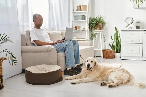 Un uomo afroamericano con miastenia gravis siede su un divano, utilizzando un computer portatile con il suo cane Labrador accanto a lui a casa.. — Foto stock