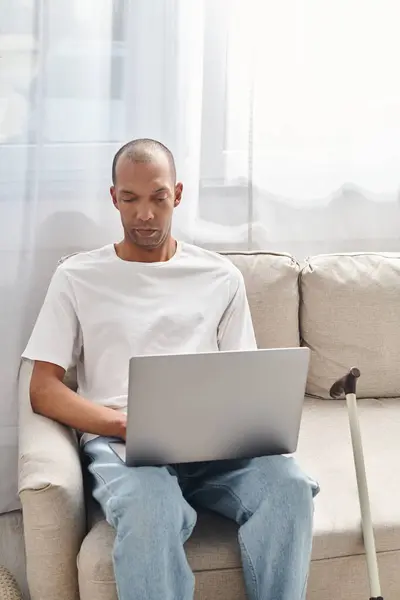 A man with myasthenia gravis uses a laptop on a couch, near walking stick — Stock Photo