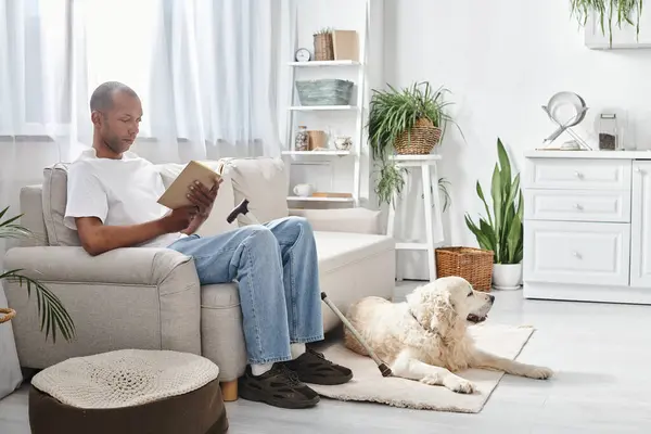Um homem afro-americano com miastenia gravis está sentado em um sofá, lendo um livro, acompanhado por seu fiel cão Labrador. — Fotografia de Stock
