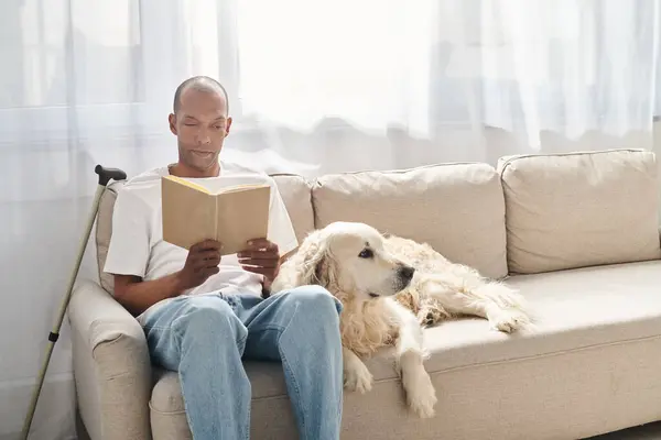 Un Afro-Américain atteint de myasthénie grave est assis sur un canapé avec son chien du Labrador, absorbé par la lecture d'un livre. — Photo de stock