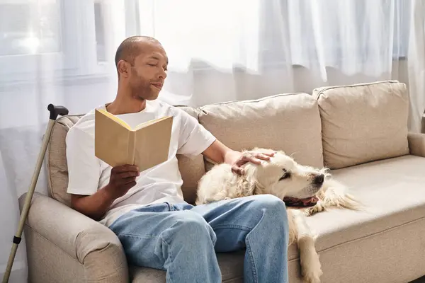 Un afroamericano discapacitado con miastenia gravis leyendo un libro en un sofá junto a su leal perro labrador. - foto de stock