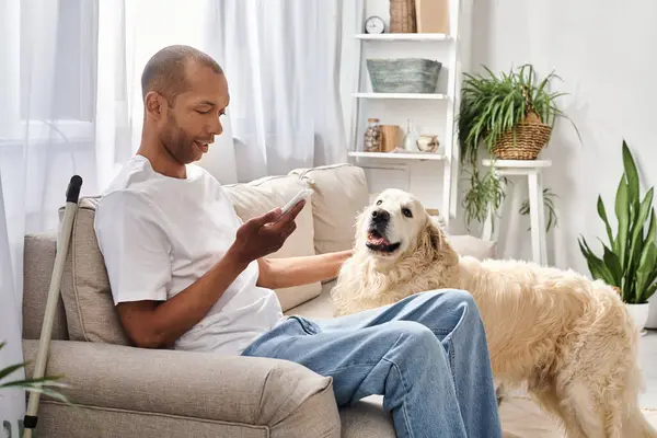 Un hombre afroamericano con miastenia gravis se sienta en un sofá, cerca de su perro Labrador y usando un teléfono inteligente - foto de stock