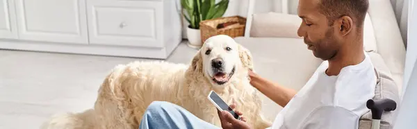 Homem afro-americano deficiente com miastenia gravis sentado em um sofá, acariciando ternamente seu cão Labrador leal. — Fotografia de Stock