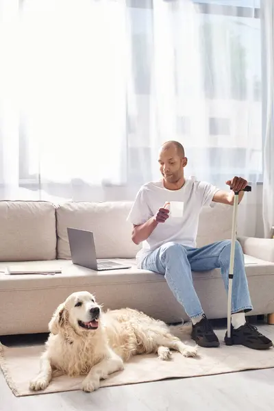 Um afro-americano deficiente com miastenia gravis sentado em um sofá ao lado de seu cão Labrador leal. — Fotografia de Stock