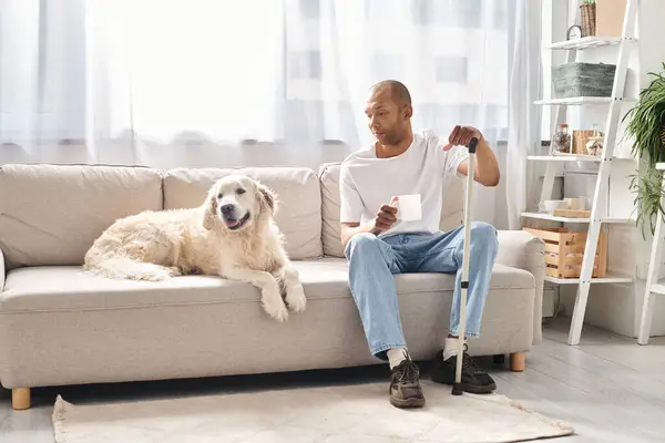 Un uomo afroamericano con miastenia gravis seduto su un divano con il suo cane Labrador al suo fianco. — Foto stock