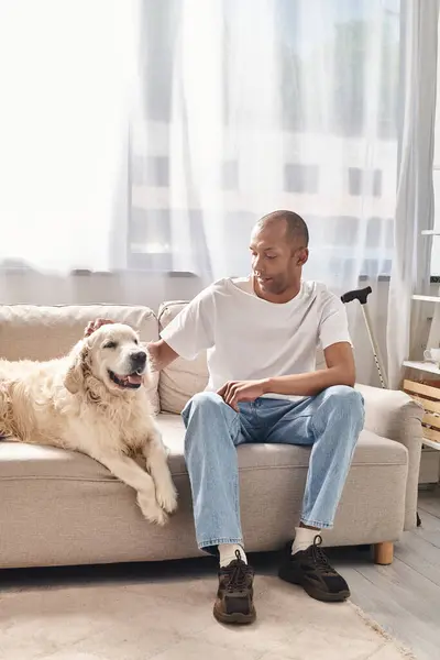 Афроамериканец с миастенией и грависом сидит на диване рядом со своей верной собакой-лабрадором в разнообразной и инклюзивной обстановке.. — стоковое фото