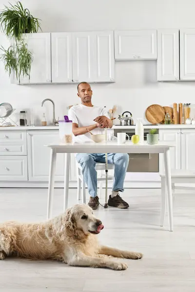 Um afro-americano deficiente senta-se em uma mesa de cozinha, acompanhado por seu cão Labrador leal. — Fotografia de Stock