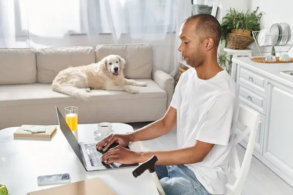 A disabled African American man sit at a table using a laptop computer, accompanied by a Labrador dog. — Stock Photo